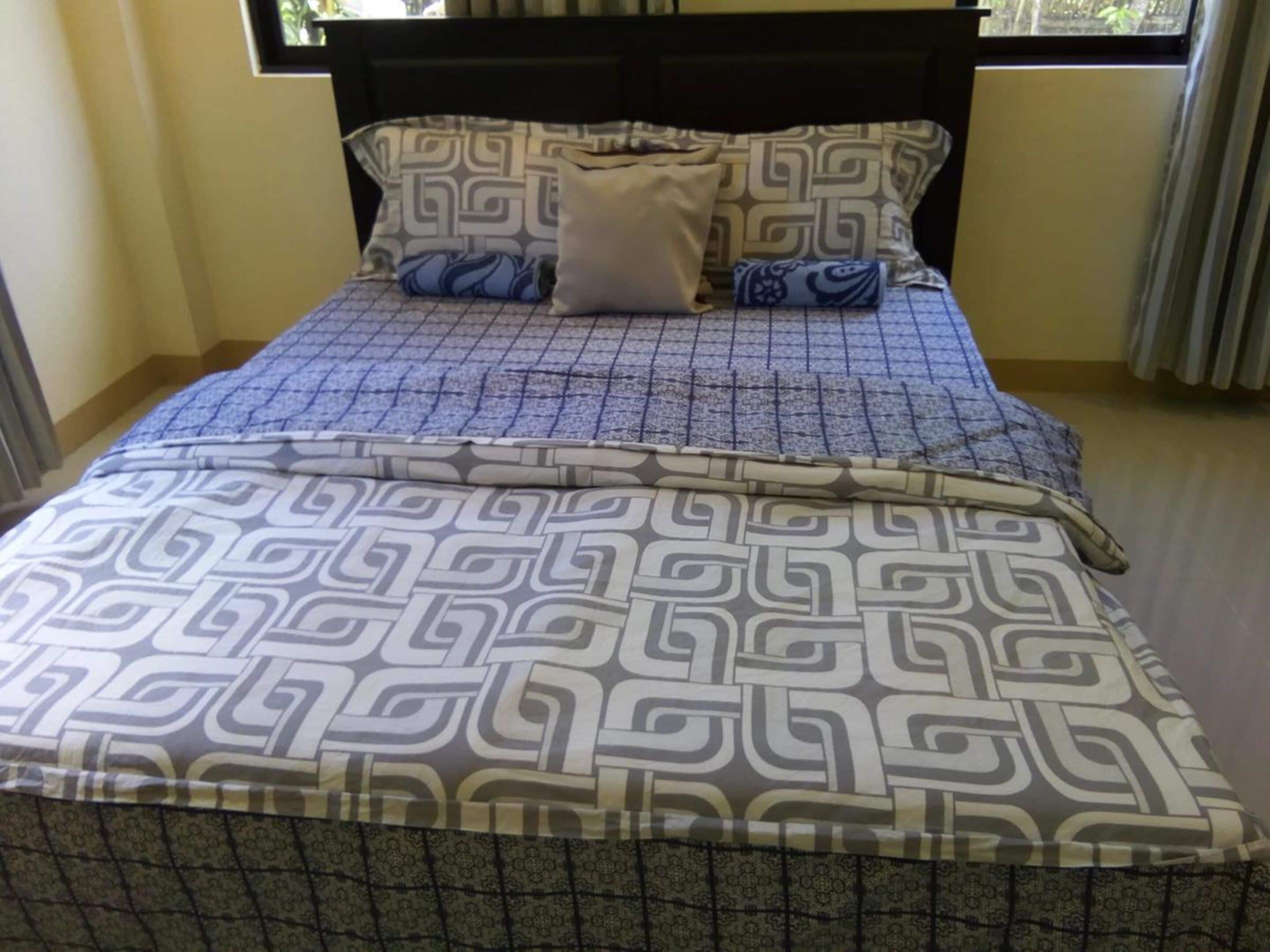 Un letto con copriletto e cuscini decorati, una testiera scura e una finestra con tende.