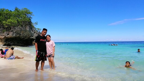 A beach scene with clear turquoise water, a rocky cliff, and people swimming and relaxing. - Bohol Sweet Home B&B (Guindulman)