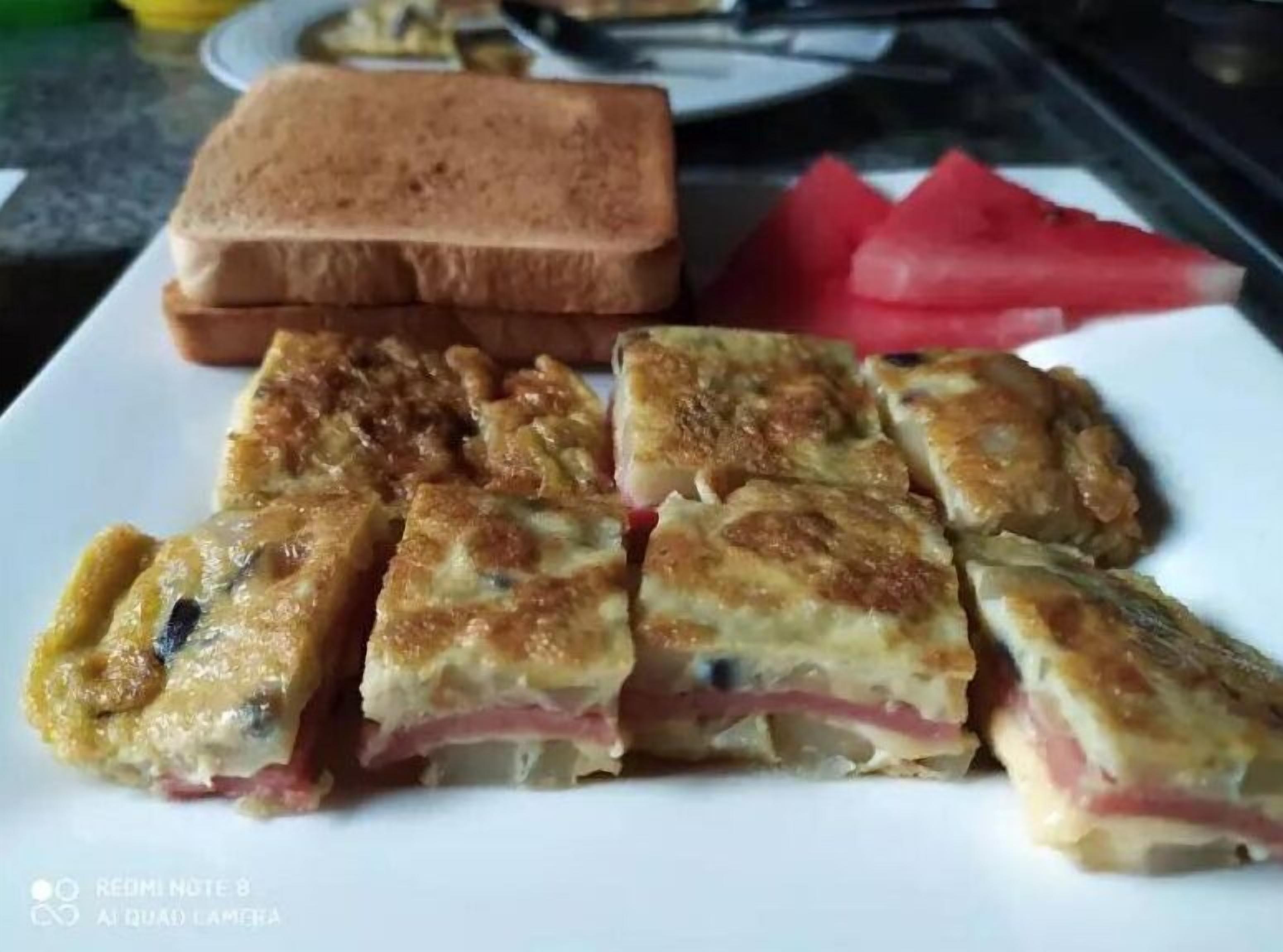 A plate with a slice of toasted bread, a slice of watermelon, and several pieces of a layered dish with cheese and ham.