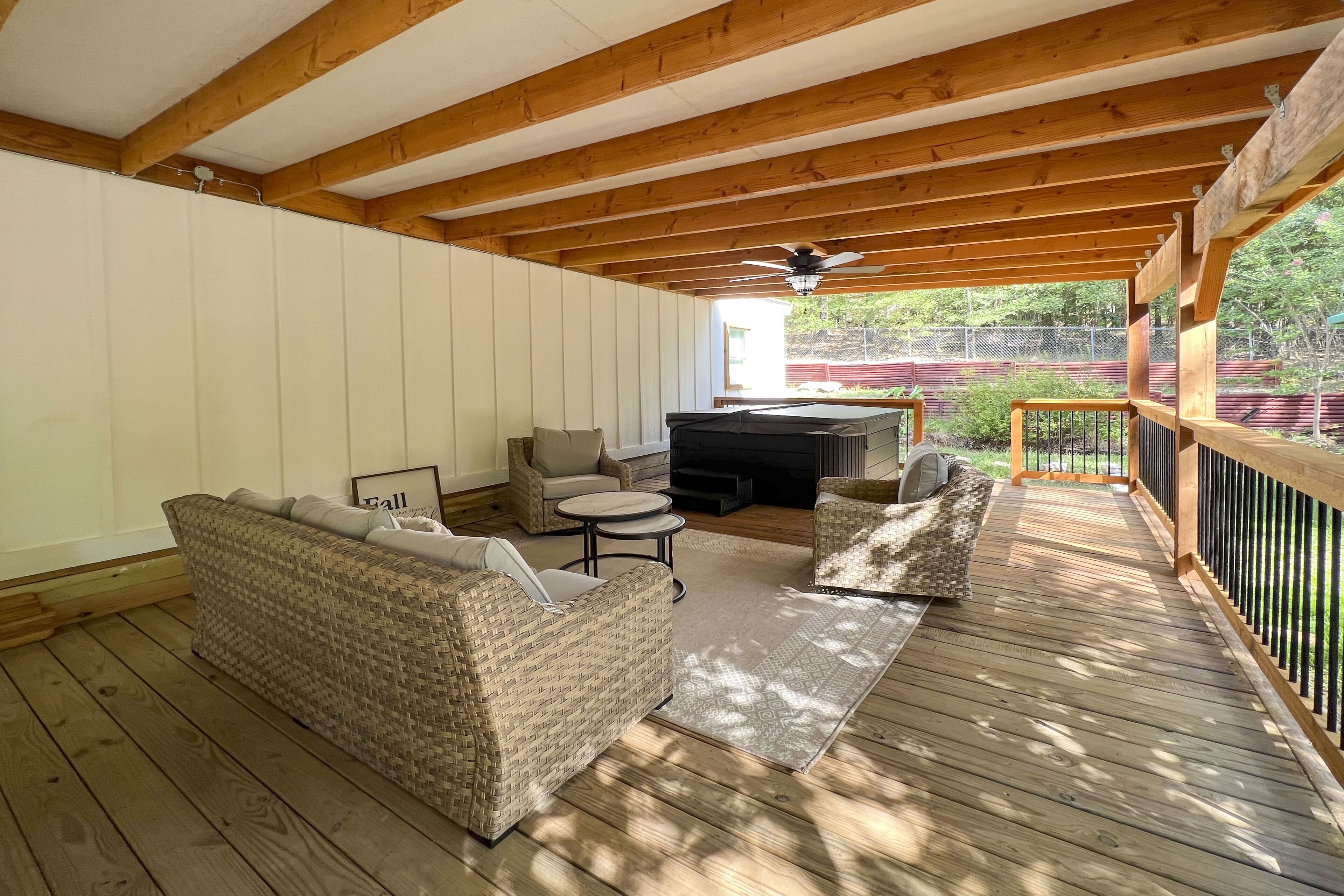 Ein überdachter Außenbereich mit Holzterrasse, einer Couch, zwei Stühlen, einem kleinen Tisch und einem Deckenventilator.