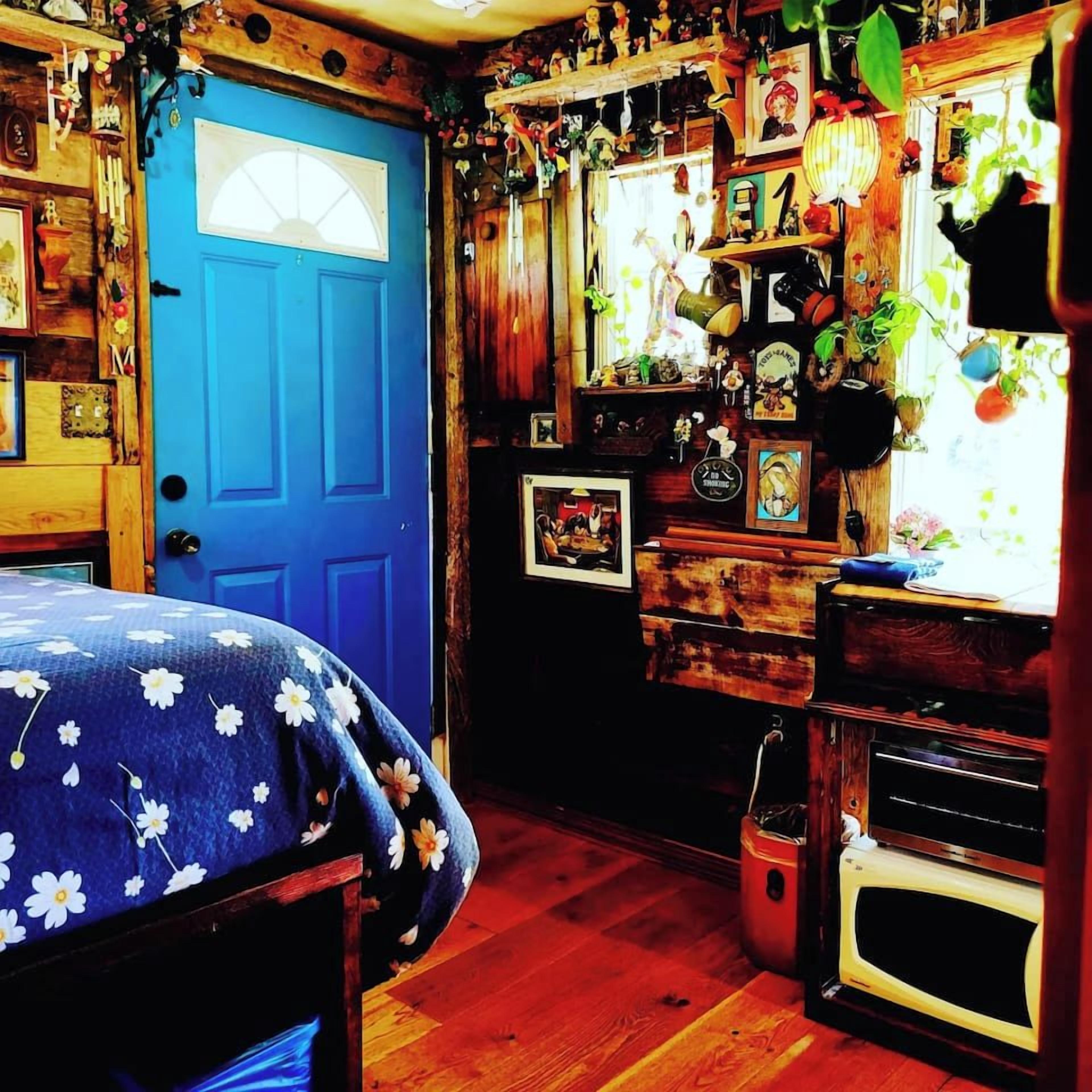 Une pièce avec une porte bleue, des murs en bois, un lit recouvert d’une couvre-lit à motifs floraux, un four à micro-ondes ancien et divers objets de décoration, notamment une horloge et des plantes suspendues.