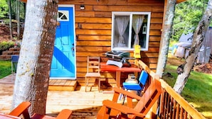 Een houten hutje met een blauwe deur, een terras met houten stoelen en tafels, en een kleine tafel met een geel voorwerp erop.