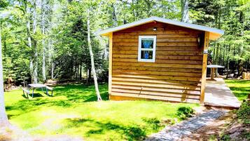 A wooden cabin with a small porch, surrounded by a grassy yard and trees.