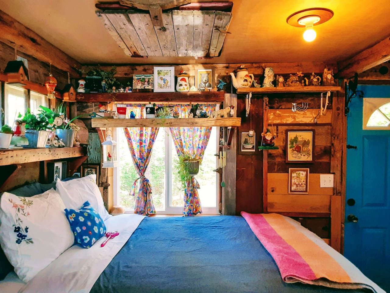 Een knus houten huisje met een bed, planken vol diverse spullen, een deur en een raam met gordijnen.