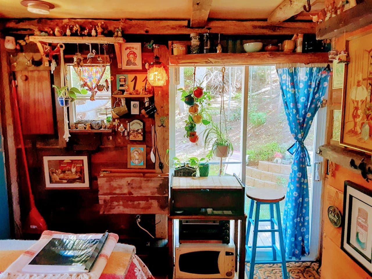 Una habitación rústica con detalles de madera, una ventana con cortinas, un taburete pequeño y varias decoraciones, incluyendo plantas y cuadros enmarcados.