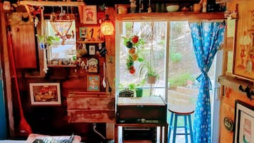 Una habitación rústica con detalles de madera, una ventana con cortinas, un taburete pequeño y varias decoraciones, incluyendo plantas y cuadros enmarcados.