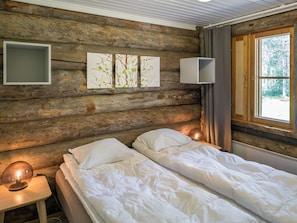 A cabin-style room with a wooden log wall, a bed with white bedding, a bedside table with a lamp, and a window with curtains.