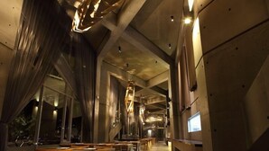 Un interior moderno con techos altos, vigas a la vista y una escalera central. Mesas y sillas están dispuestas a lo largo de las paredes, con amplios ventanales que proporcionan luz natural.