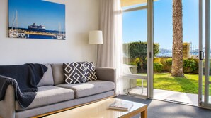 A modern hotel room with a sofa, a coffee table, a lamp, and a large window offering a view of the sea and greenery. - Wake up to sweeping harbour views in this modern, split-level apartment located between Fishing Boat Harbour and Challenger Harbour. Whether you're a couple, a family, or here on business, this peaceful retreat offers the best of coastal charm and ci (Fremantle)