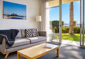 A modern hotel room with a sofa, a coffee table, a lamp, and a large window offering a view of the sea and greenery.
