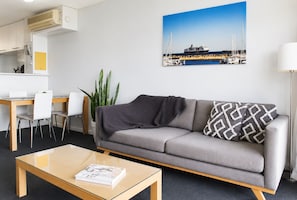 A modern living room with a sofa, a coffee table, a dining area with chairs, a potted plant, and a painting of a marina.