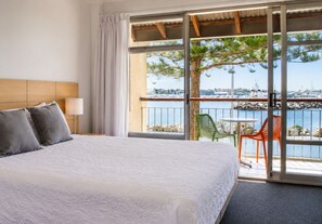 A hotel room with a bed, a nightstand, a lamp, a balcony with a table and chairs, and a view of the marina.