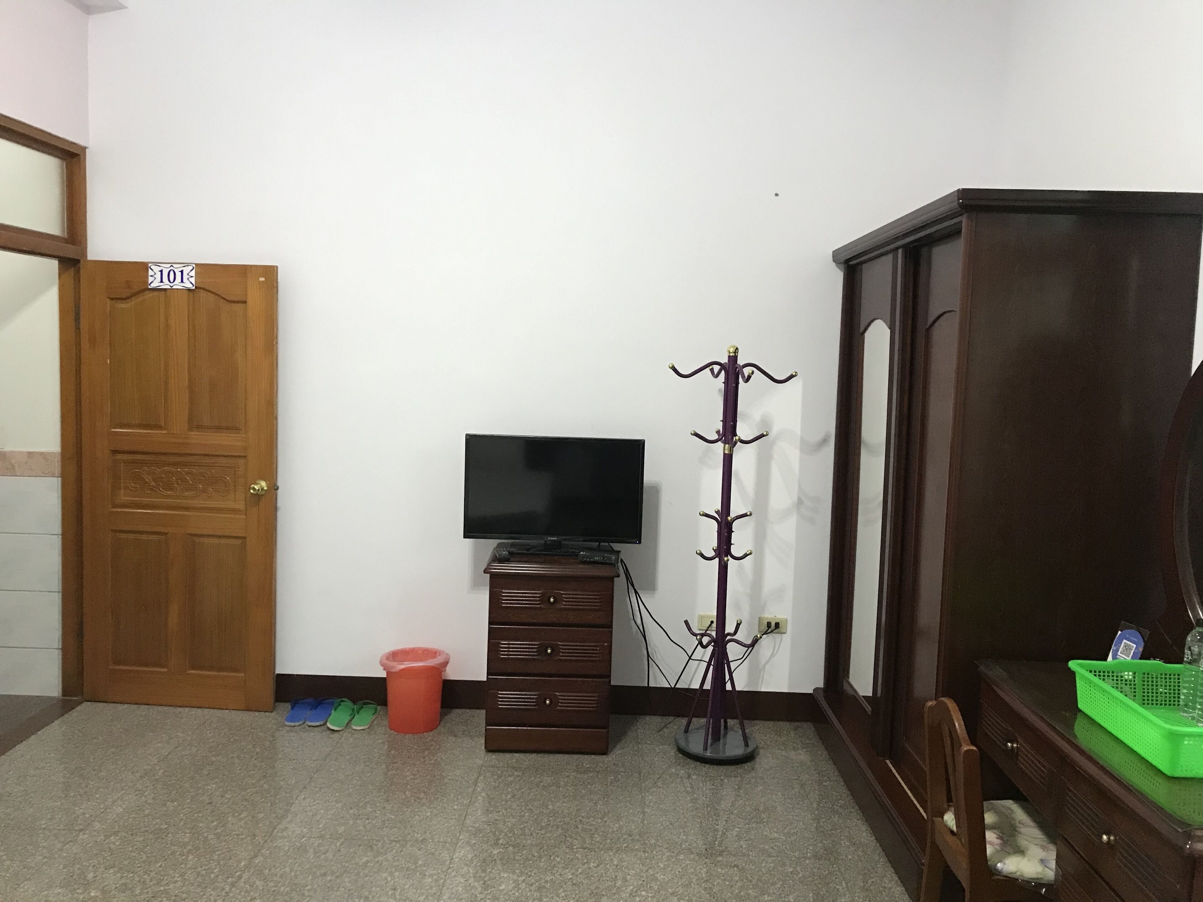 Une pièce avec une porte en bois portant l’inscription « 101 », un téléviseur posé sur un meuble en bois, un porte-manteau, une commode et un panier vert sur la commode.