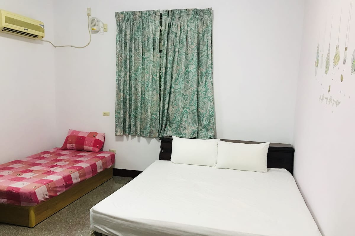 A bedroom with a single bed, a smaller bed, a wall-mounted air conditioner, and a window with patterned curtains.