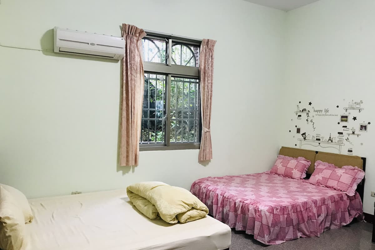 A bedroom with a bed, a window with curtains, a wall decoration, and a bed with a pink bedspread.