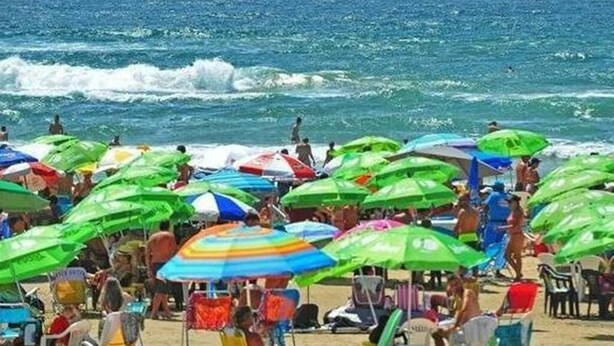 Ein überfüllter Strand mit vielen bunten Sonnenschirmen und Menschen, die das Meer genießen.