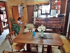 Uma sala de jantar com mesa e cadeiras de madeira, um vaso com flores, e uma cozinha ao fundo.