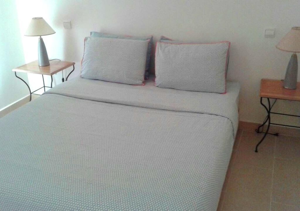 Un lit bien fait, avec une couverture grise et deux oreillers, une table de chevet avec une lampe et une prise électrique au mur.