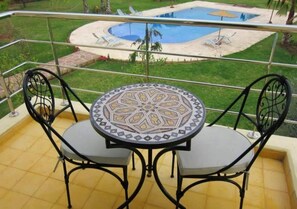 Ein runder Tisch mit einer gemusterten Platte und zwei Stühle mit Kissen, auf einem Balkon mit Blick auf einen Swimmingpool und Liegestühle.