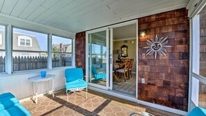 A covered patio with blue furniture, a wooden wall, and a sliding glass door leading to an interior dining area. - Oceanside Beach House in Pvt Beach Community! (Mantoloking)