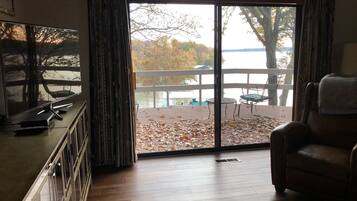 A room with a large sliding glass door leading to a deck with a view of a lake and trees.