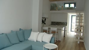 Een moderne woonkamer met een blauwe bank, een klein wit tafeltje en een keuken met witte kasten en een eettafel.