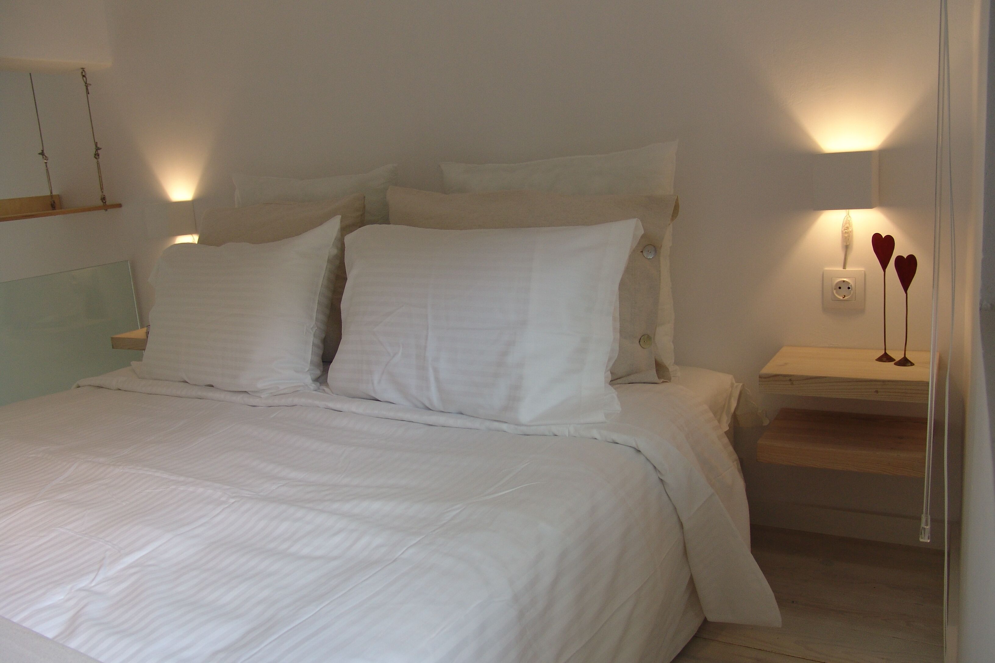 Ein ordentlich bezogenes Bett mit weißen Leinen, ein Nachttisch mit Lampe und eine Steckdose an der Wand.
