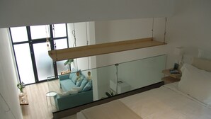 Een moderne slaapkamer met een groot bed, een glazen scheiding en uitzicht op een zithoek met een bank en een plant.