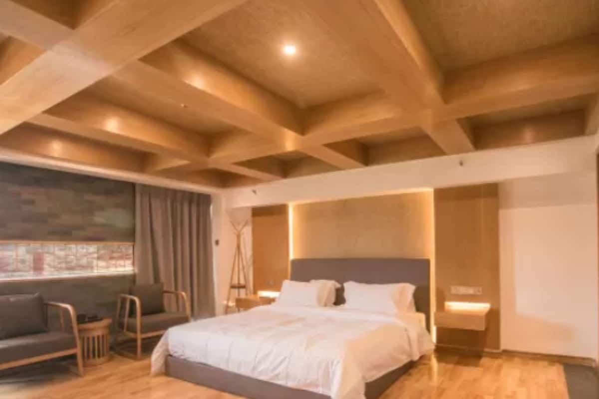 A modern bedroom with a wooden ceiling, a large bed, a seating area with a sofa and chairs, and a view through a window.