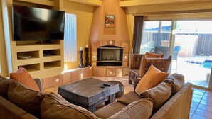 A living room with a fireplace, a television, and a pool visible through the glass door. - Serene Desert Escape-Salt Water Pool & Jacuzzi/Fire Pit/Views #067047 3bd (La Quinta)