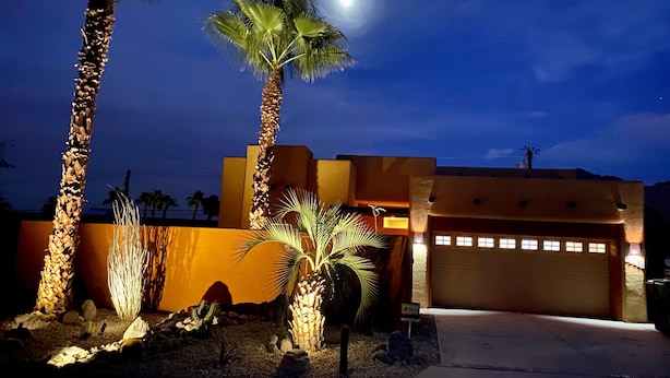 A modern house with a garage, palm trees, and a well-lit driveway at night. - Serene Desert Escape-Salt Water Pool & Jacuzzi/Fire Pit/Views #067047 3bd (La Quinta)