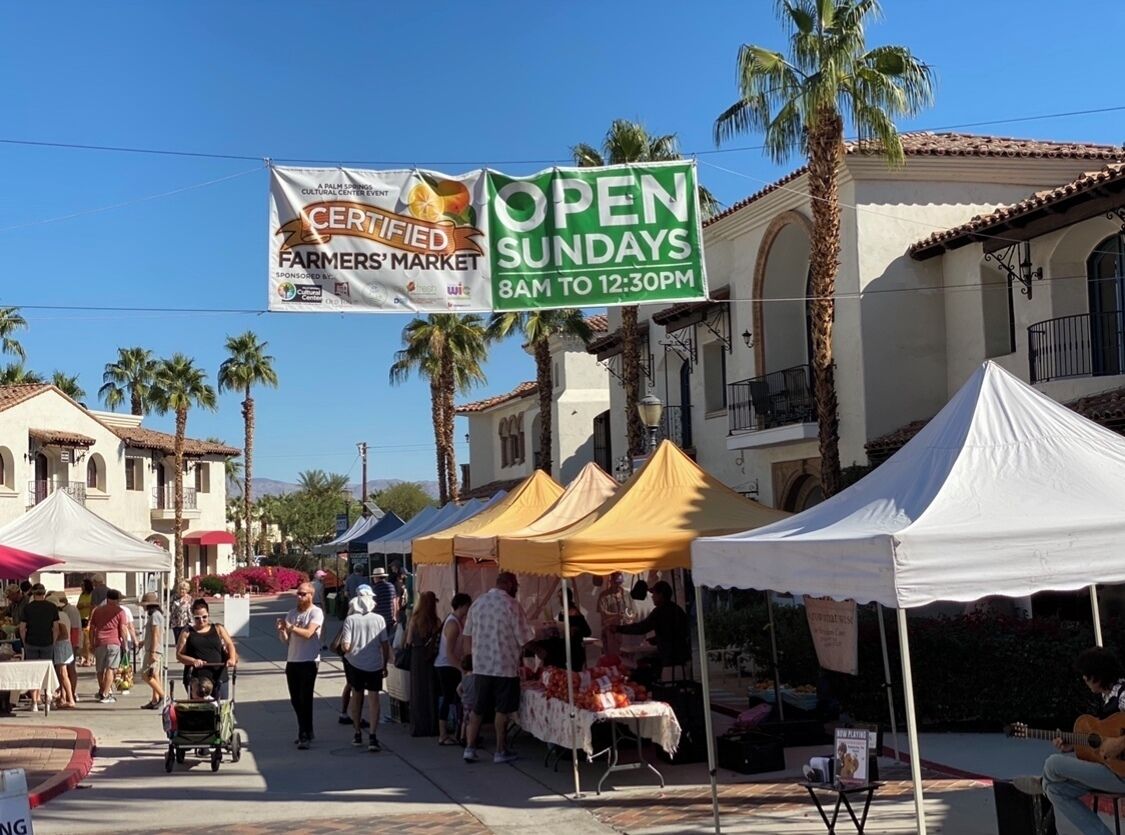A farmers' market with tents and a sign indicating open Sundays from 8 AM to 12:30 PM.