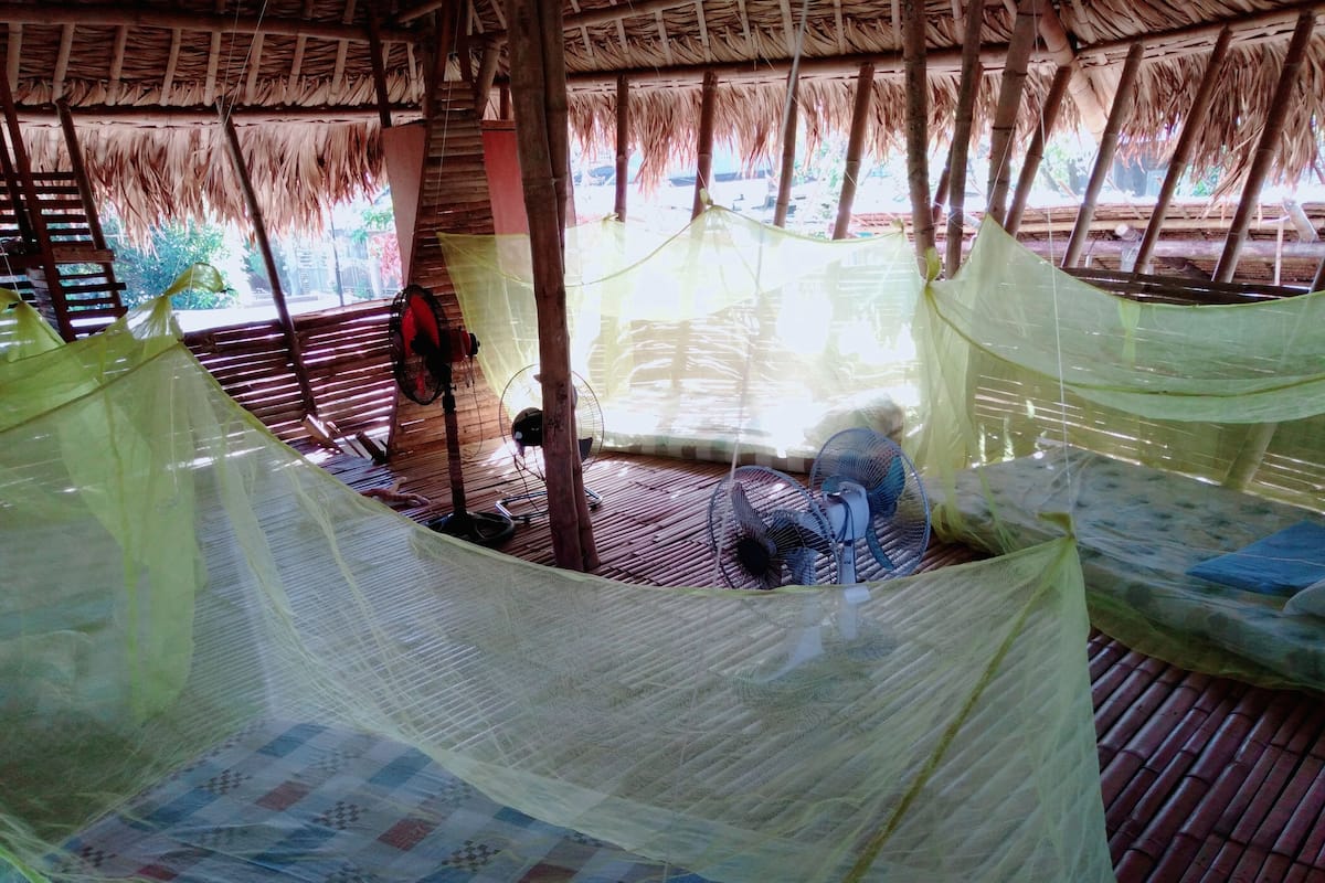 A thatched-roof room with a bed, a fan, and a mosquito net.