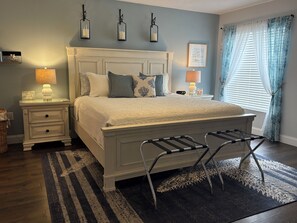 A bedroom with a bed, nightstands, a lamp, a wall-mounted shelf, a window with curtains, and a hanging lamp. - Captain's Cove Waterside View Condo 103, walk to the beach or enjoy the pool! (Indian Shores)