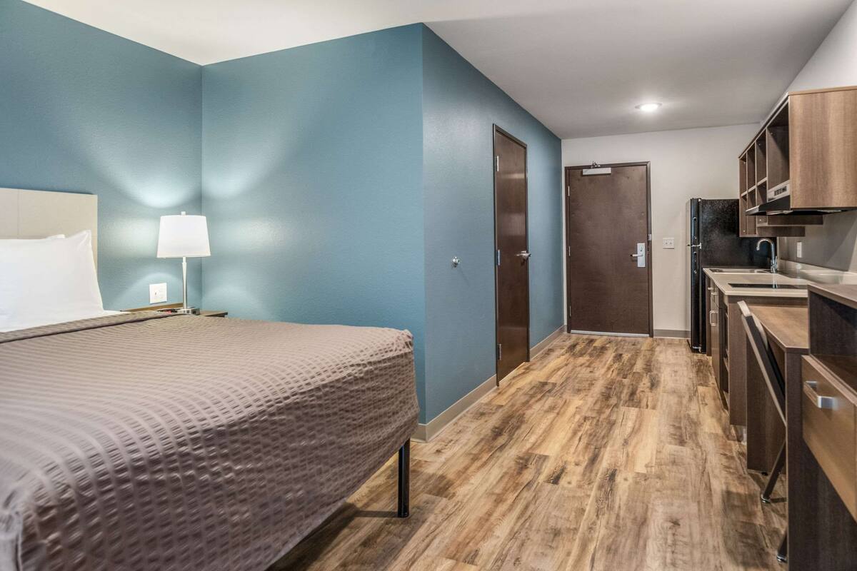 A hotel room with a bed, a nightstand, a lamp, a wooden floor, a kitchenette, and a door.