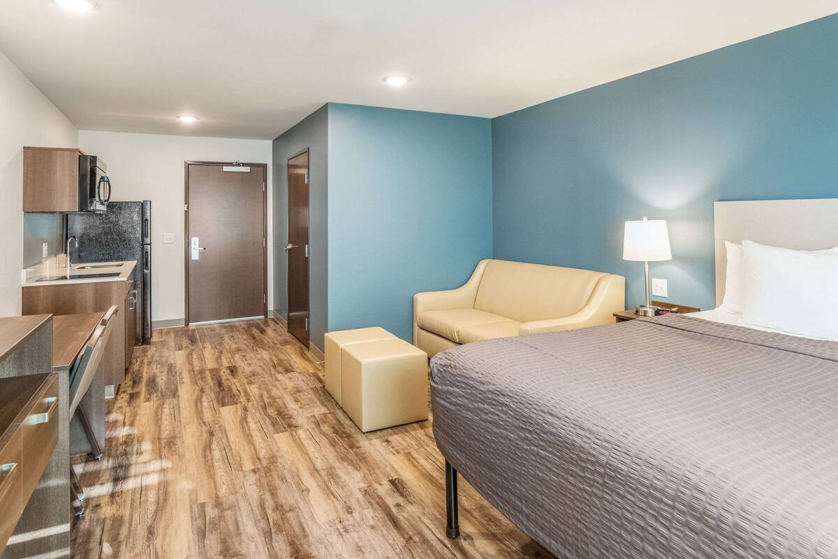A hotel room with a bed, a sofa, a desk with a computer, a kitchenette, and a door leading to another room.