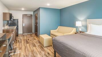 A hotel room with a bed, a sofa, a desk with a computer, a kitchenette, and a door leading to another room.