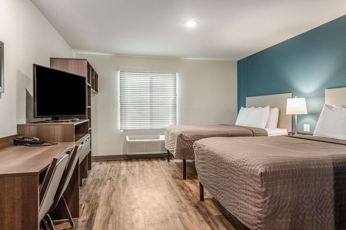 A hotel room with a desk, two beds, a TV, and a window with blinds.
