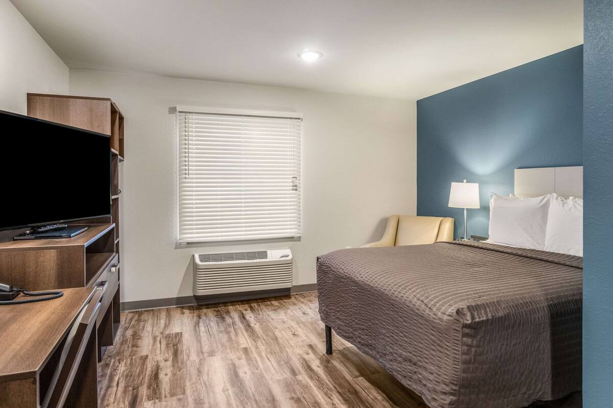 A hotel room with a bed, a desk with a TV, a lamp, and a window with blinds.