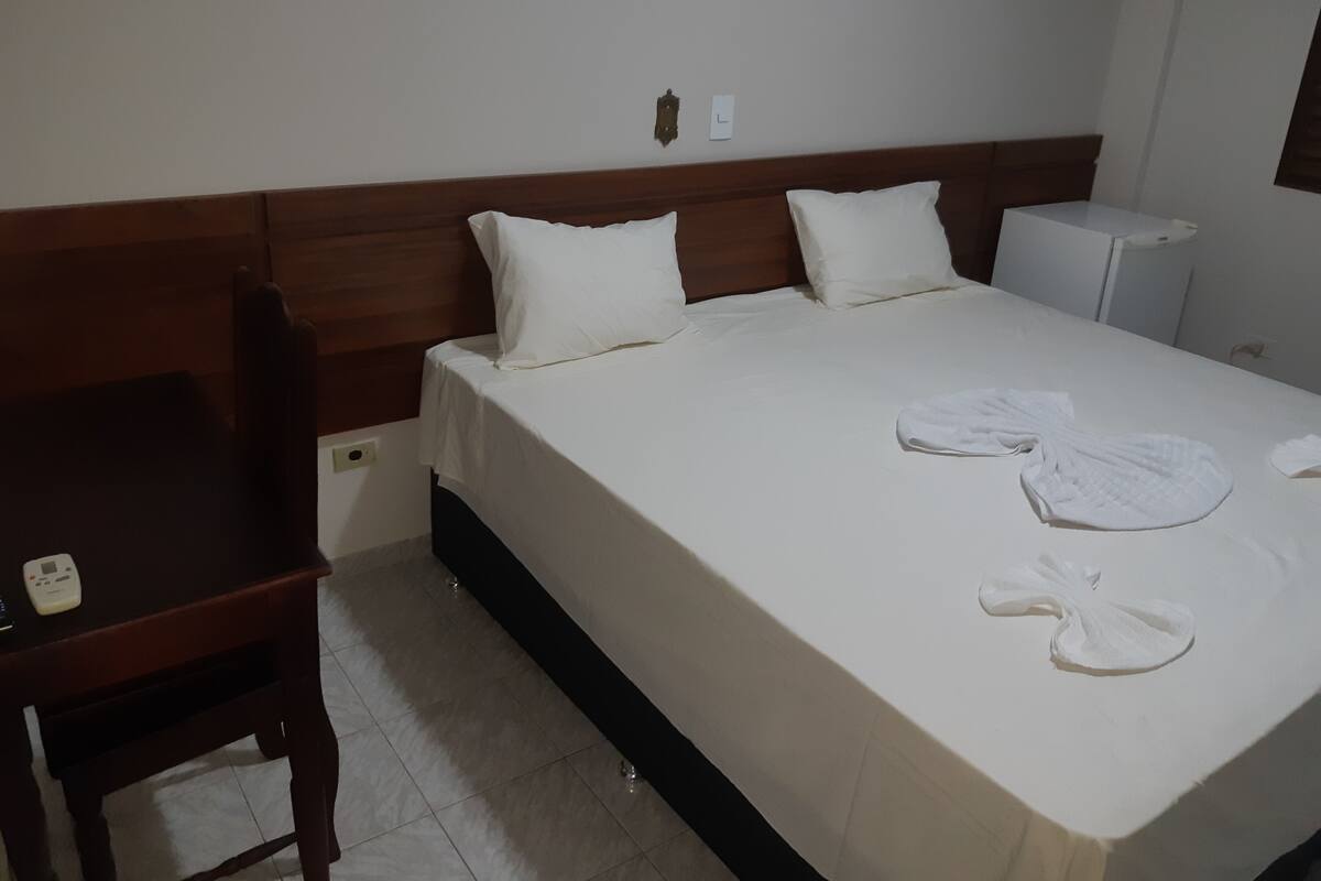 A neatly made bed with white linens and two pillows, a wooden nightstand with a lamp, and a small desk with a telephone.