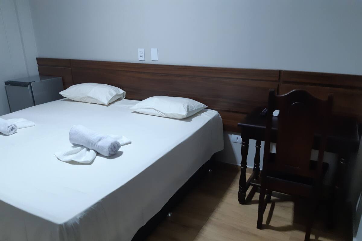 A neatly made bed with white linens and pillows, a wooden headboard, a small bedside table, and a chair.