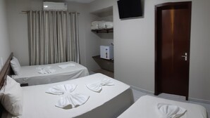 Habitación de hotel con dos camas, cada una con una toalla doblada en forma de lazo. Se observa un refrigerador pequeño y un estante con toallas dobladas. También se ve una puerta de madera y un televisor montado en la pared.