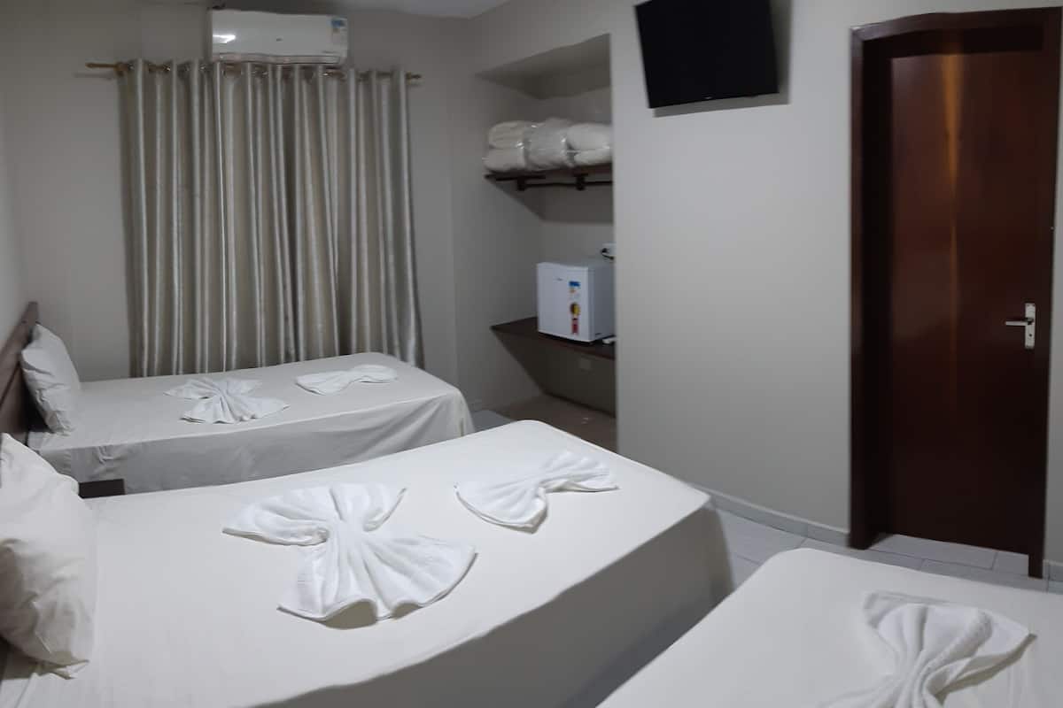 A hotel room with two beds, each with a towel arranged in a bow shape. There is a small refrigerator and a shelf with folded towels. A wooden door and a wall-mounted TV are also visible.