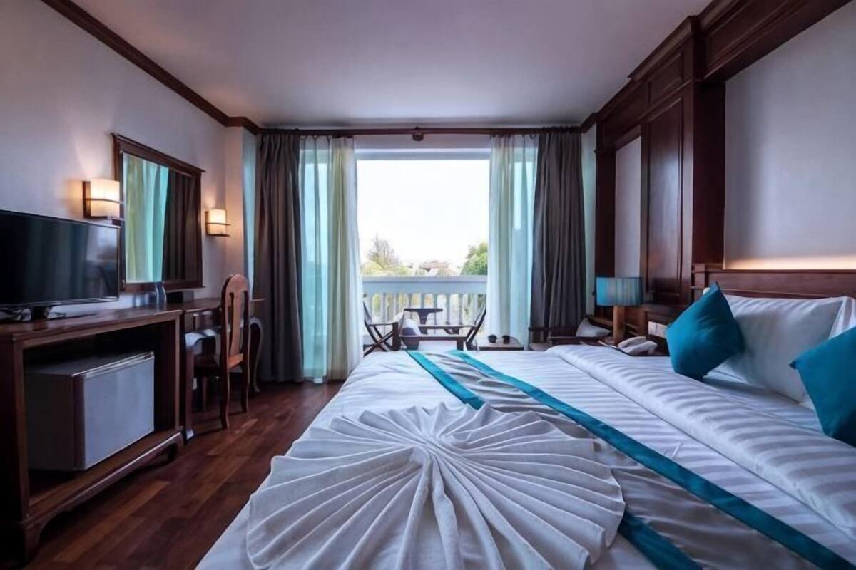 Una camera d'albergo con un letto, una scrivania con televisione, una sedia, un balcone con vista e una finestra con tende.