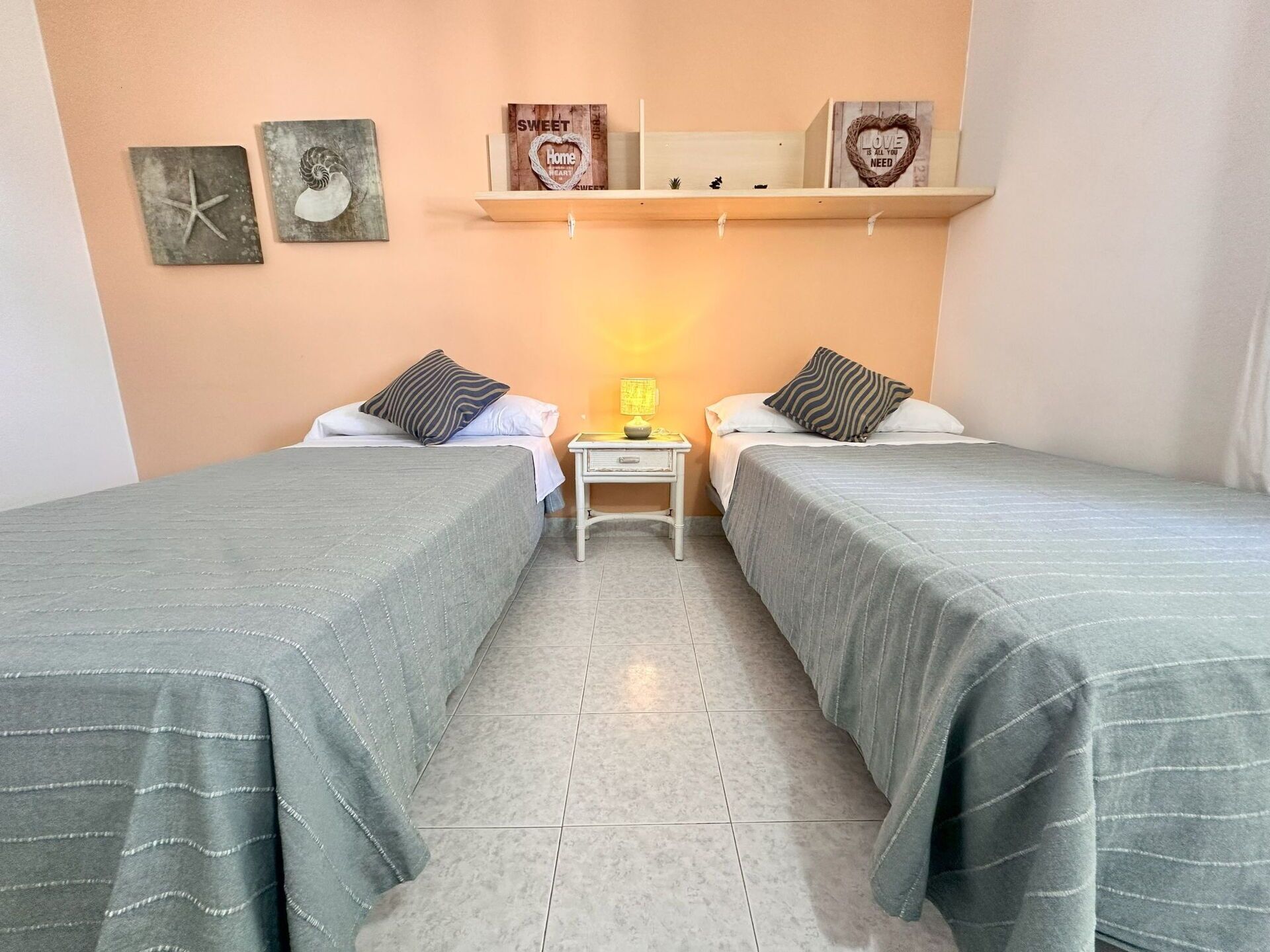A twin room with two beds, a nightstand, a shelf with decorative items, and a wall with framed pictures.