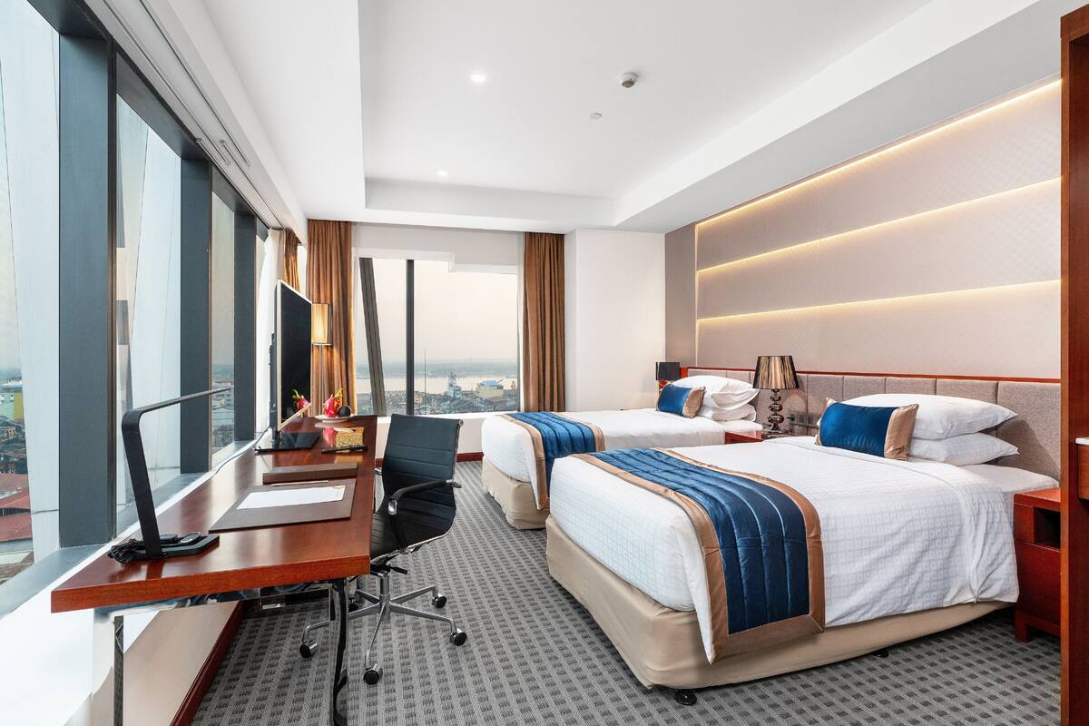 A hotel room with two beds, a desk with a chair, a large window, and a city view.