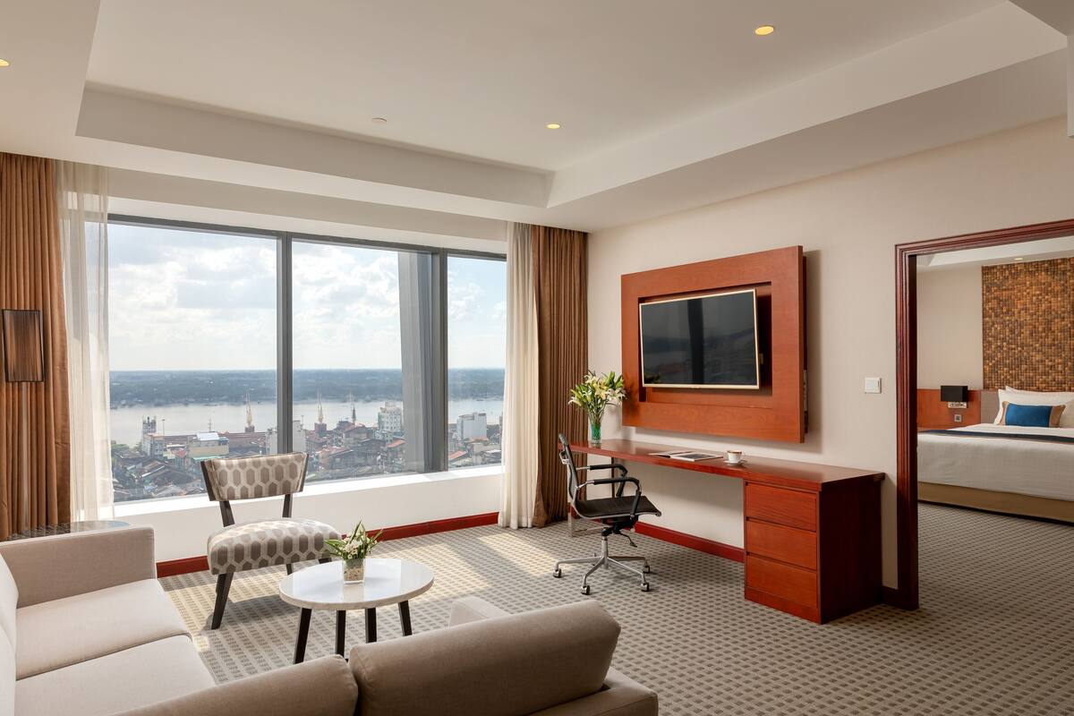 A hotel room with a large window, a desk, a chair, a TV, and a bed.