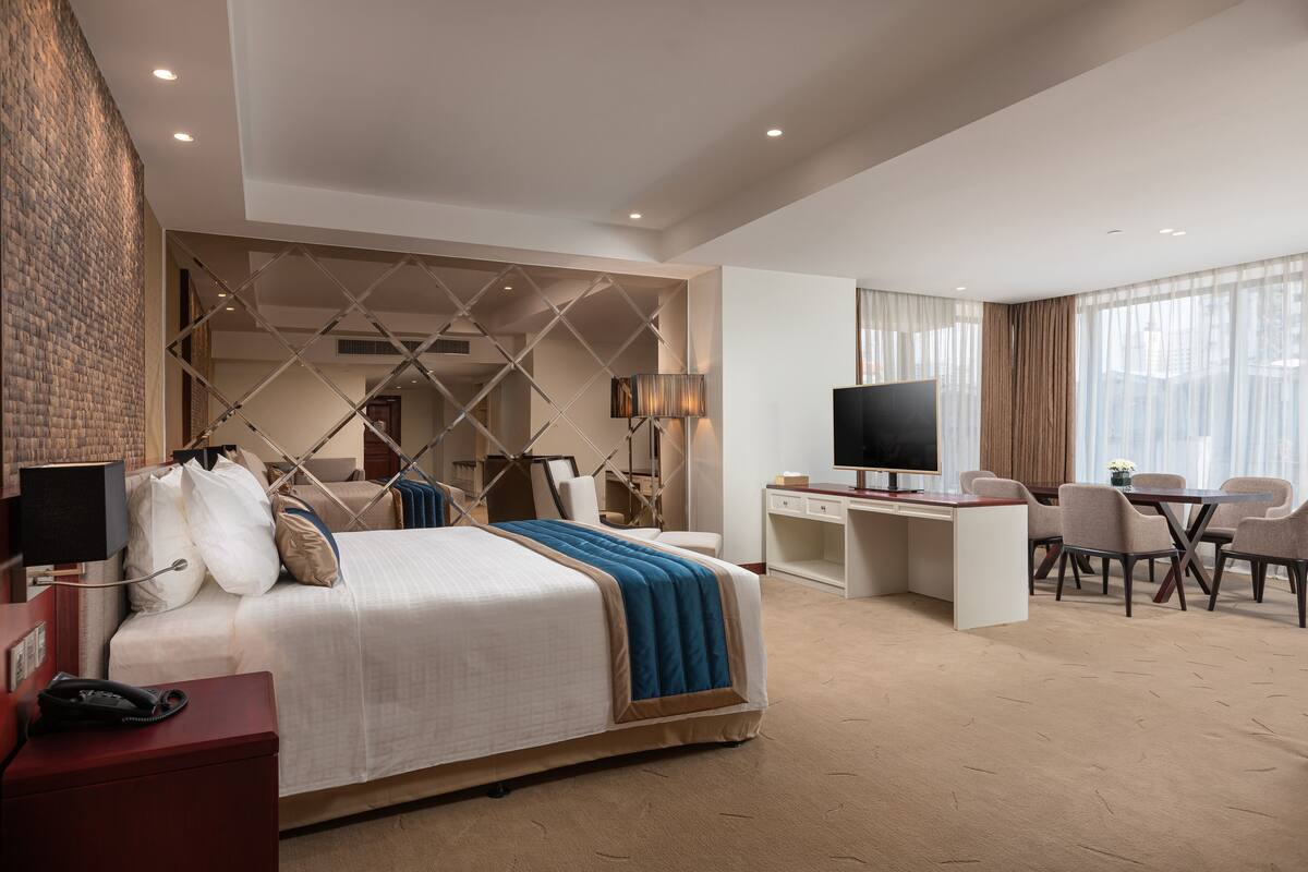 A hotel room with a large bed, a desk, a TV, and a dining area with chairs.