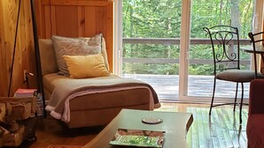 Ein gemütliches Zimmer mit Holzboden, einer Couch mit Kissen, einem Tisch mit einem Gesellschaftsspiel und einem großen Fenster mit Blick ins Grüne.