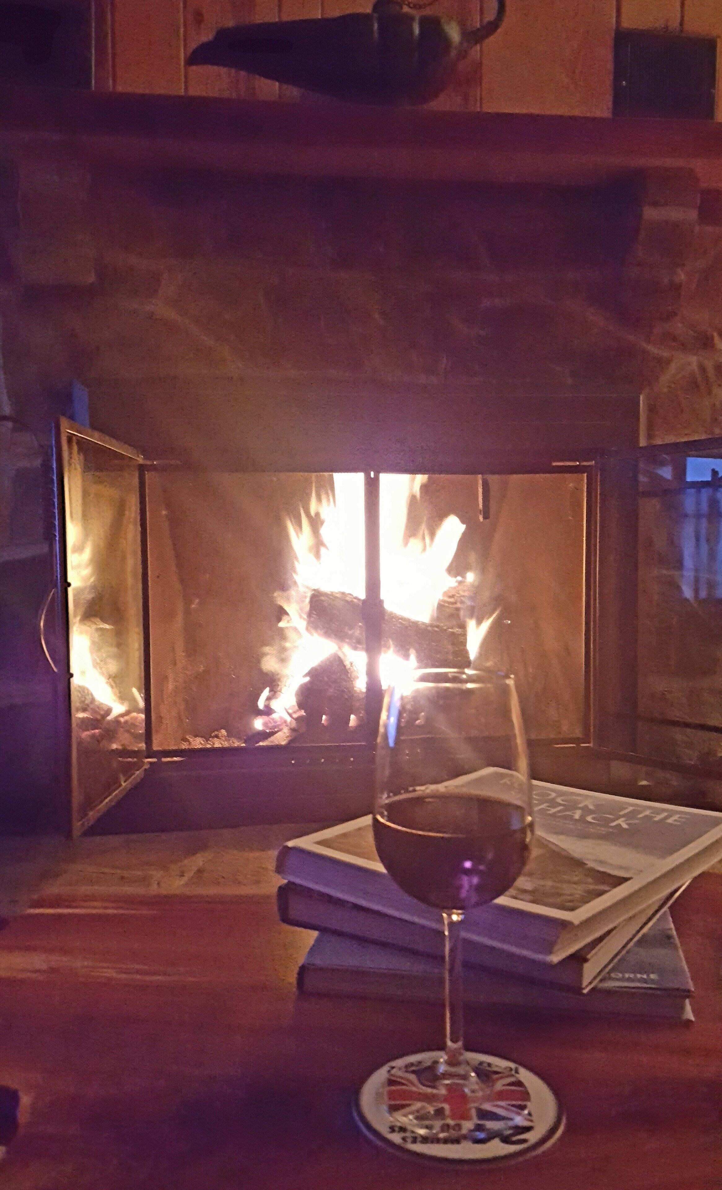 A glass of red wine on a coaster with a book and a magazine on a wooden table in front of a fireplace with a visible fire.